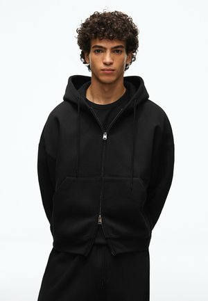 Jeune homme aux cheveux bouclés portant un sweat à capuche noir zippé et un pantalon noir, debout devant un fond blanc uni.