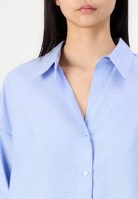 Šviesiai mėlyna marškinėlių tipo palaidinė su apykakle, lygi tekstūra, laisvas kirpimas ir išskirtinės siūlės detalių išdėstymas prie sagų.