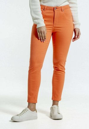 Jeans en denim orange avec une coupe slim, présentant une fermeture à bouton et à zip, des poches avant et des ourlets retroussés, associés à des baskets blanches.