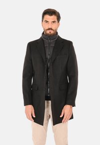 Uomo con barba curata che indossa un cappotto nero sopra una giacca nera con zip e pantaloni beige, in piedi davanti a uno sfondo chiaro e uniforme.