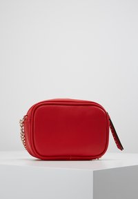 Punainen keinonahkainen laukku suorakulmaisella muodolla, ketjuhihnalla ja korostetulla vetoketjulla. Sileä pinta, minimalistinen muotoilu ilman näkyviä kuvioita.