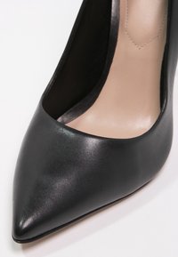 Svart läder damspetsig pumpsko med beige innersula, visad i närbild mot vit bakgrund.