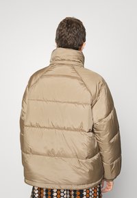 Beige pufferjakke med høj krave, vandret quiltning og elastiske manchetter. Glat, shiny tekstur; har en afslappet pasform.