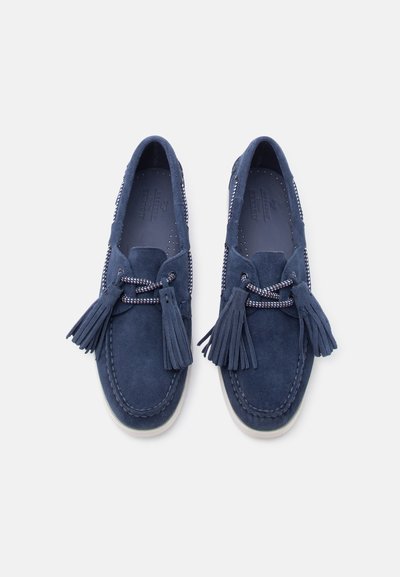 Ζευγάρι μοκασίνια από σουέτ σε ναυτικό μπλε με λευκή σόλα, πλεκτά κορδόνια και μεγάλες φούντες, σε άνωθεν θέαση σε λευκό φόντο.