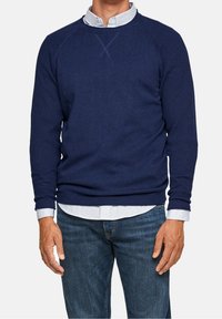 Homme portant un pull à manches longues bleu marine sur une chemise blanche à motifs, associé à un jean bleu, debout devant un fond uni.