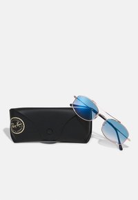 Ray-Ban'i prillid, millel on sinise gradatsiooniga objektiivid ja kerge kuldne metallraam, puhkavad mustal tekstureeritud karbil, millel on logo.