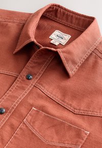 Brick-red button-up shirt with a textured fabric, featuring a point collar, white stitching, and a chest pocket. Round black buttons included.