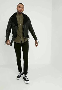 Uomo in piedi che indossa una giacca di pelle nera con collo di pelliccia, una camicia verde militare con bottoni, pantaloni neri e sneakers nere con suola bianca.