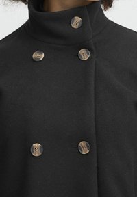 Černý vlněný kabát s vysokým límcem, dvojitým zapínáním a pěti kulatými krémovými knoflíky s tmavými akcenty.