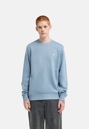 Giovane uomo con capelli rossi corti che indossa una felpa azzurro chiaro e jeans grigio scuro, in piedi davanti a uno sfondo bianco semplice.
