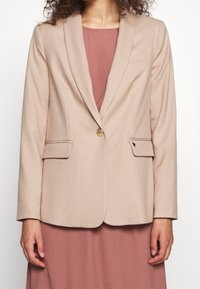 Blazer beige chiaro con alette, bottone d'oro singolo, due tasche frontali e una texture liscia. Indossato su un vestito rosa.