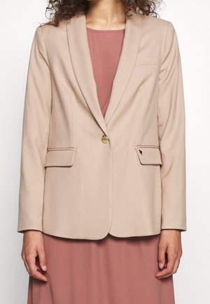 Blazer beige clair avec des revers, un bouton doré unique, deux poches avant et une texture lisse. Porté sur une robe rose.