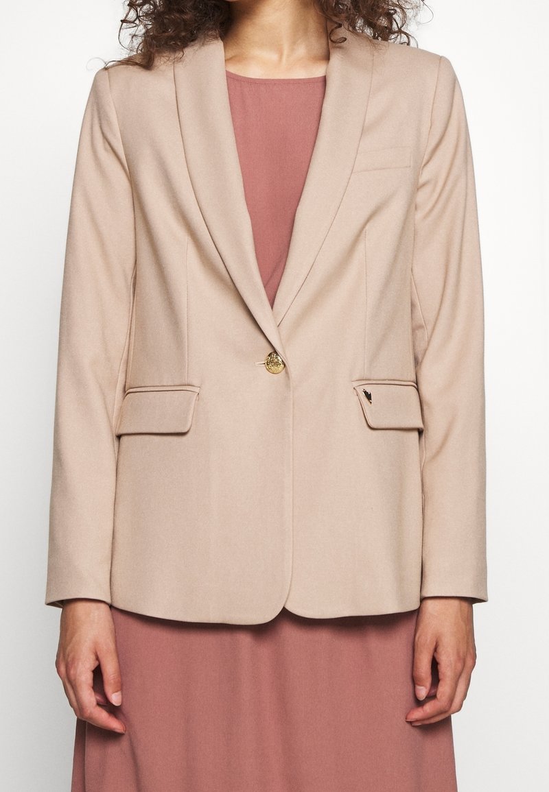 Blazer beige chiaro con alette, bottone d'oro singolo, due tasche frontali e una texture liscia. Indossato su un vestito rosa.