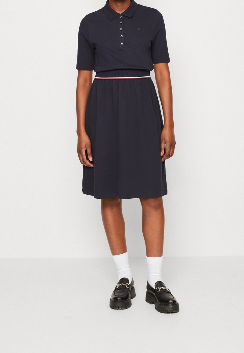 Personne portant une robe polo bleu marine à manches courtes, des chaussettes blanches et des mocassins noirs épais, debout devant un fond blanc uni.