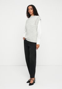 Sivý sveter s rebrovanými detailmi, nosený cez bielu košeľu, v kombinácii s čiernymi vysokými džínami a čiernymi lodičkami, stojaci na bielom pozadí.