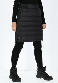 Svart isolerad kjol med quiltat mönster, justerbar midja, två sidofickor med dragkedja och logodetalj vid fållen, matchad med svarta leggings och stövlar.