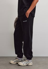 Persona che indossa pantaloni della tuta Pegador neri, maglietta nera, sneakers bianche, in piedi con una mano in tasca davanti a un muro semplice.