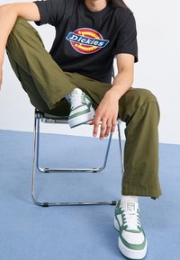 Czarna koszulka z logo "Dickies", oliwkowe spodnie i białe trampki z zielonymi akcentami. Model siedzi na chromowanym krześle na niebieskim tle.