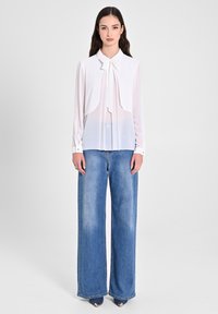 Camicia bianca con fiocco, design a strati e polsini con bottone, abbinata a jeans in denim blu a gamba larga con una texture sbiadita.