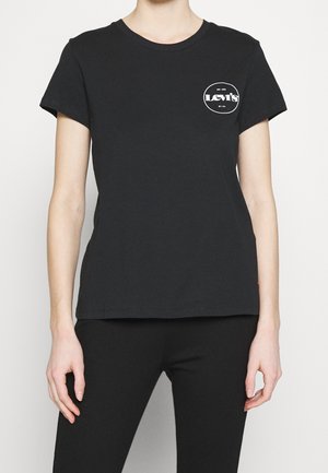 Personne portant un T-shirt noir Levi's avec un petit logo blanc sur la partie supérieure gauche de la poitrine, associé à un pantalon noir, debout sur un fond blanc.