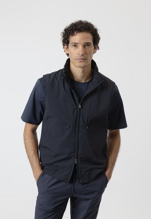 Hombre con cabello rizado y oscuro lleva una camisa azul marino de manga corta, chaleco sin mangas con cremallera y pantalones a juego, de pie con las manos en los bolsillos.