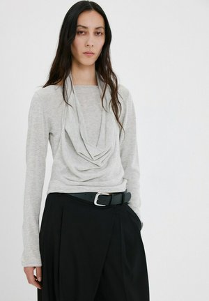 Mujer con cabello largo y oscuro que lleva una blusa gris claro con cuello drapeado metida en pantalones negros de talle alto con cinturón negro.