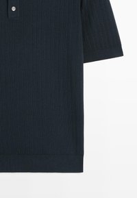 Polo à manches courtes en maille côtelée bleu marine foncé avec deux boutons blancs sur la patte de boutonnage, visible de la poitrine à l'ourlet sur un fond blanc.