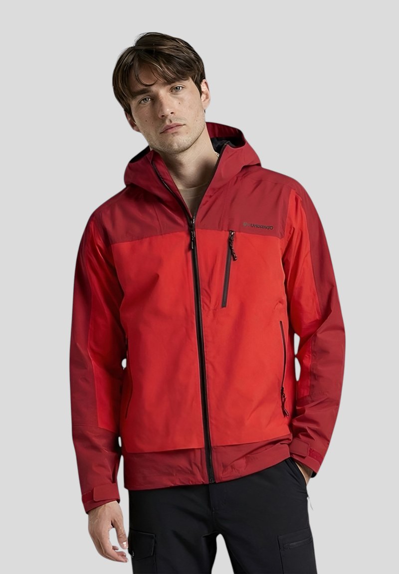 Junger Mann trägt eine rote Outdoorjacke mit Kapuze, schwarzen Reißverschlüssen und schwarzen Hosen, steht vor einem schlichten grauen Hintergrund.