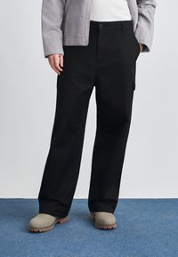 Pantaloni cargo negri din bumbac, cu croial�ă lejeră, buzunare laterale și închidere cu nasturi, asortați cu pantofi slip-on gri pe o suprafață albastră.