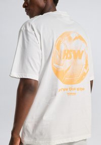 Hvid bomulds T-shirt med et stort orange fodboldgrafik og teksten "HSW" og "serve the game" på ryggen. Korte ærmer, afslappet pasform.