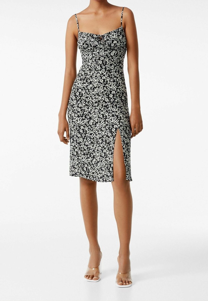 Abito midi nero a fiori con spalline sottili, corpetto aderente e spacco laterale. Il tessuto presenta un motivo floreale bianco su tutta la superficie, con una texture liscia.