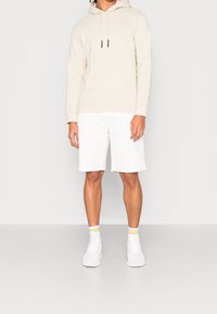 Sweat à capuche beige avec une poche avant, associé à un short blanc. La tenue est complétée par des baskets blanches et des chaussettes montantes rayées.