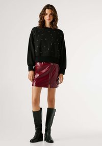 Melns sporta krekls ar apaļiem acuļiem, valkāts kopā ar spīdīgu burgundijas ādas mini svārku ar sānu rāvējslēdzēju. Melni zābaki līdz ceļiem papildina izskatu.