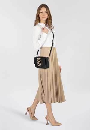 Borsa a tracolla nera con lettering dorato "LOVE", materiale liscio, forma rettangolare, tracolla regolabile; indossata con una blusa bianca e una gonna plissettata beige.