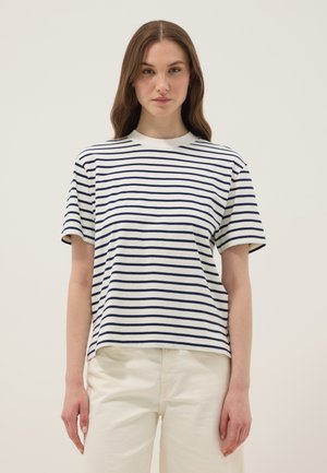 Frau mit langen braunen Haaren, die ein weiß-navy gestreiftes Kurzarmshirt und helle Hose vor einem neutralen Hintergrund trägt.