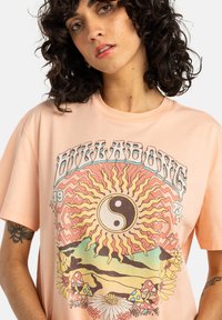 Pfirsichfarbiges Baumwoll-T-Shirt mit einem grafischen Design, das eine Sonne, das Yin-Yang-Symbol, Pilze und Blumen in lebendigen Farben zeigt. Kurze Ärmel, lockere Passform.