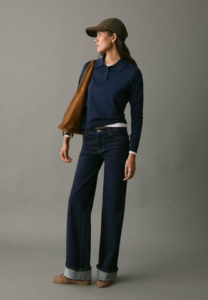 Suéter de punto navy con dobladillo acanalado, combinado con jeans de mezclilla oscuros de campana y un bolso de hombro marrón. Una gorra de cuadros marrón completa el atuendo.