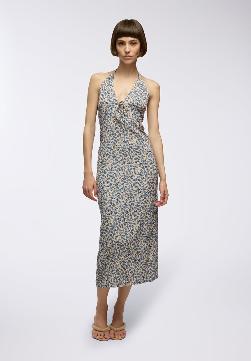 Abito a halter floreale in crema con motivi di fiori blu, corpino aderente e lunga gonna. La modella indossa sandali beige.