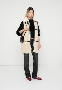 Beige faux shearling väst med svarta accenter, svart tröja, mörka jeans och röda skor. Dragkedjestängning och sidofickor. Liten handväska.