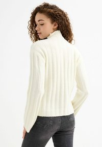 Suéter acanalado crema con cuello alto y mangas largas y acampanadas, confeccionado en un suave material texturizado. La vista posterior muestra una silueta ajustada.