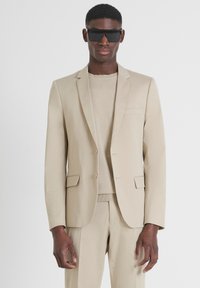 Completo beige su misura con giacca monopetto, rever a punta e due tasche frontali. Indossato sopra un maglione leggero coordinato.