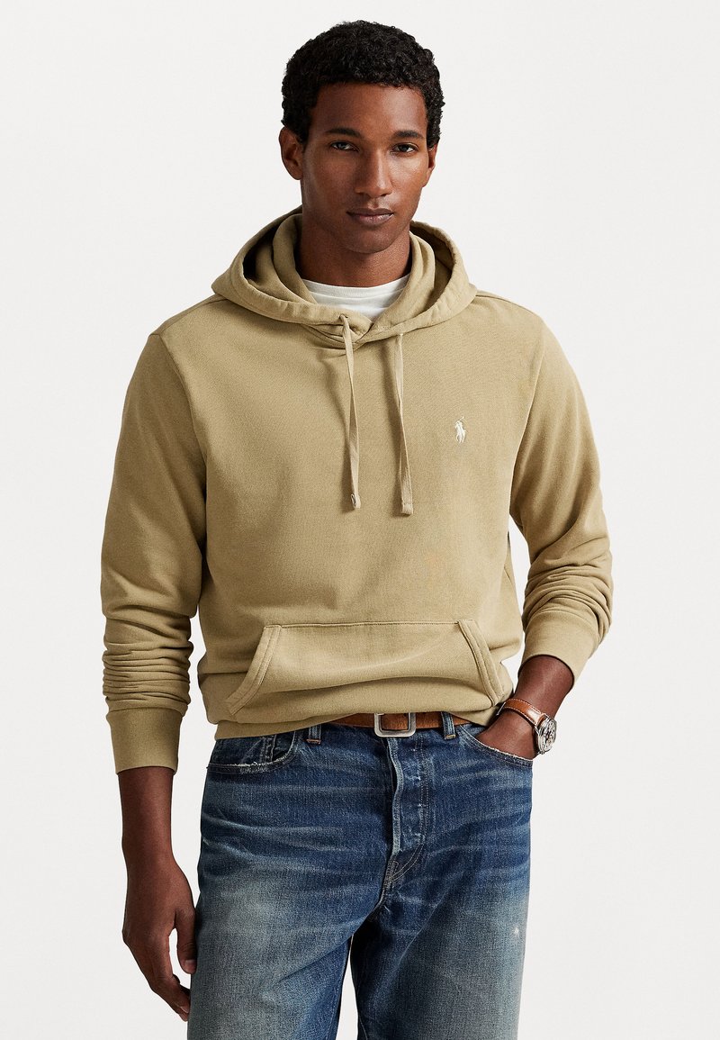 Man in een beige hoodie met een voorzak en blauwe jeans, staand met één hand in de zak tegen een witte achtergrond.