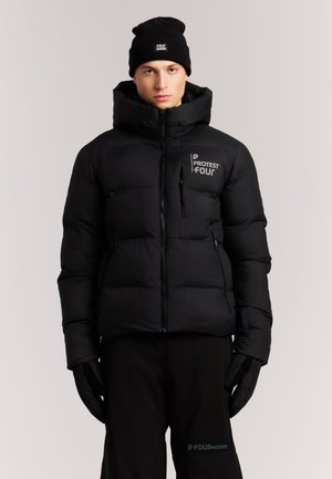 Jong persoon draagt een zwarte winterjas, zwarte muts, handschoenen en broek met "Protest Four" logo's op jas en broek.