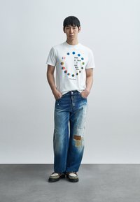 Man standing with hands in pockets, wearing white t-shirt with colorful circle design and text, blue patched jeans, and green-white shoes.