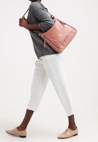 Sac à main en cuir rose clair avec une finition texturée, doté de sangles réglables et d'une poche zippée. Porté par une personne en pantalon blanc.