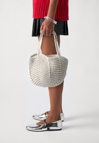 White, woven tote bag with a rounded shape and two short handles. Textured surface with a crochet pattern. Flat silver shoes worn.
