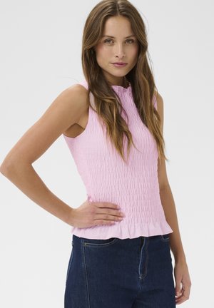 Pruunide pikkade juustega naine, kes kannab varrukateta roosat lokkis topi ja tumesiniseid teksaseid, poseerides ühe käega puusal.