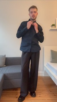 Zwarte overhemdd met lange mouwen, gecombineerd met brede zwarte broeken. Het shirt heeft een kraag en twee borstzakken.