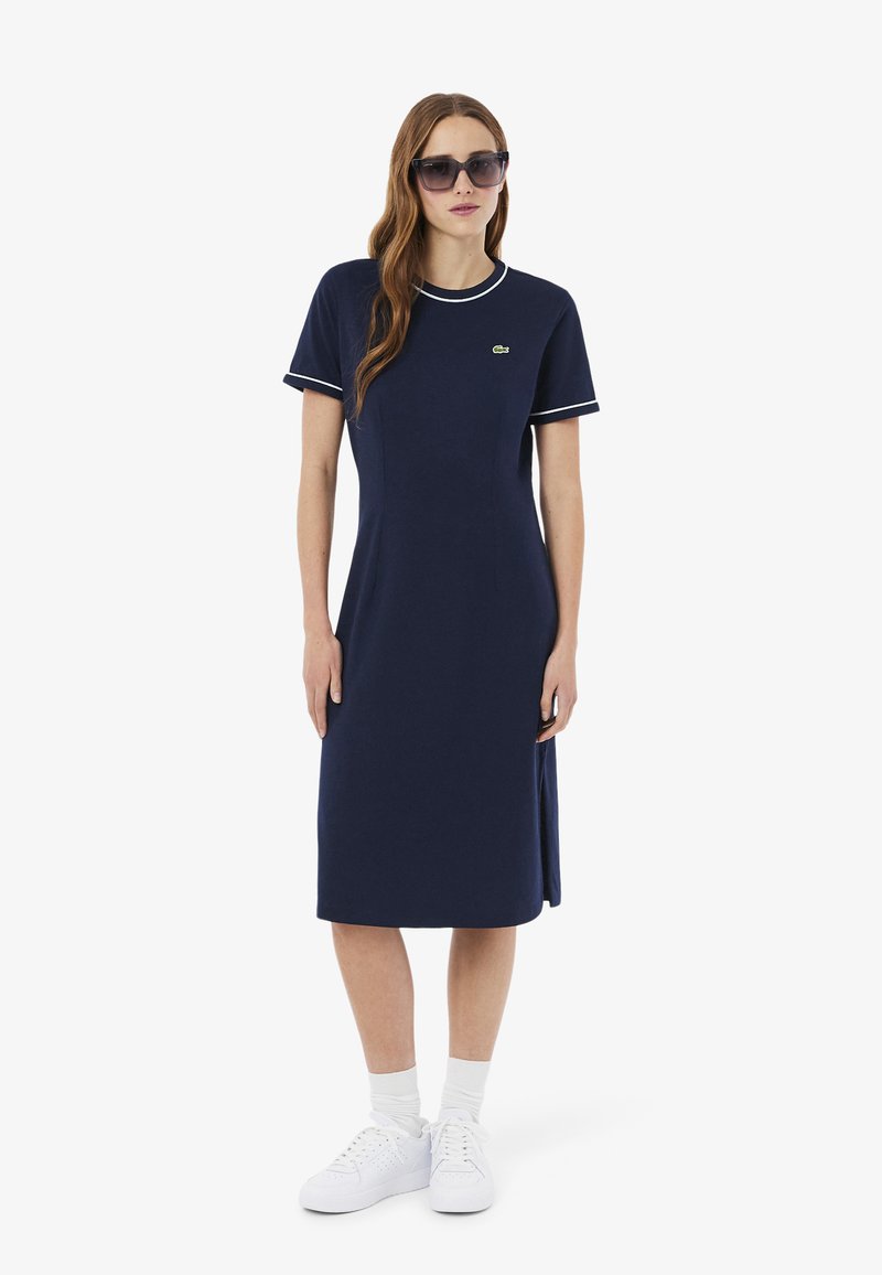 Vrouw draagt een marineblauwe midi-jurk met witte bies aan mouwen en kraag, witte sokken, witte sneakers en grote donkere zonnebril.