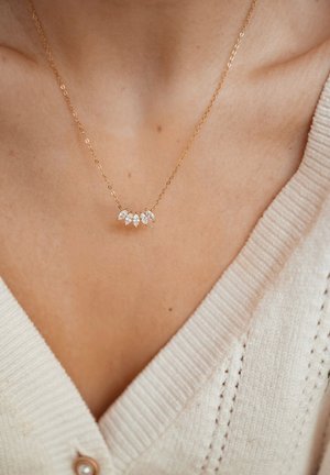 Delicate gouden ketting met vijf marquise-geslepen heldere edelstenen, gedragen op een blote hals boven een crèmekleurige V-hals trui.